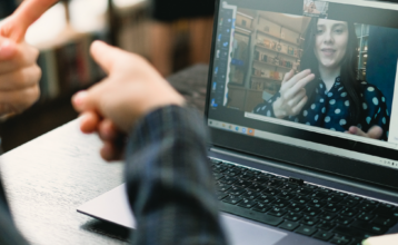 Über die Schulter einer nicht zu identifizierenden Person sieht man dessen Hände mit ausgestreckten Zeigefingern. Vor ihr steht ein Laptop. Sie unterhält sich in Gebärdensprache mit einer weiblich gelesenen Person auf dem Bildschirm des Laptops.