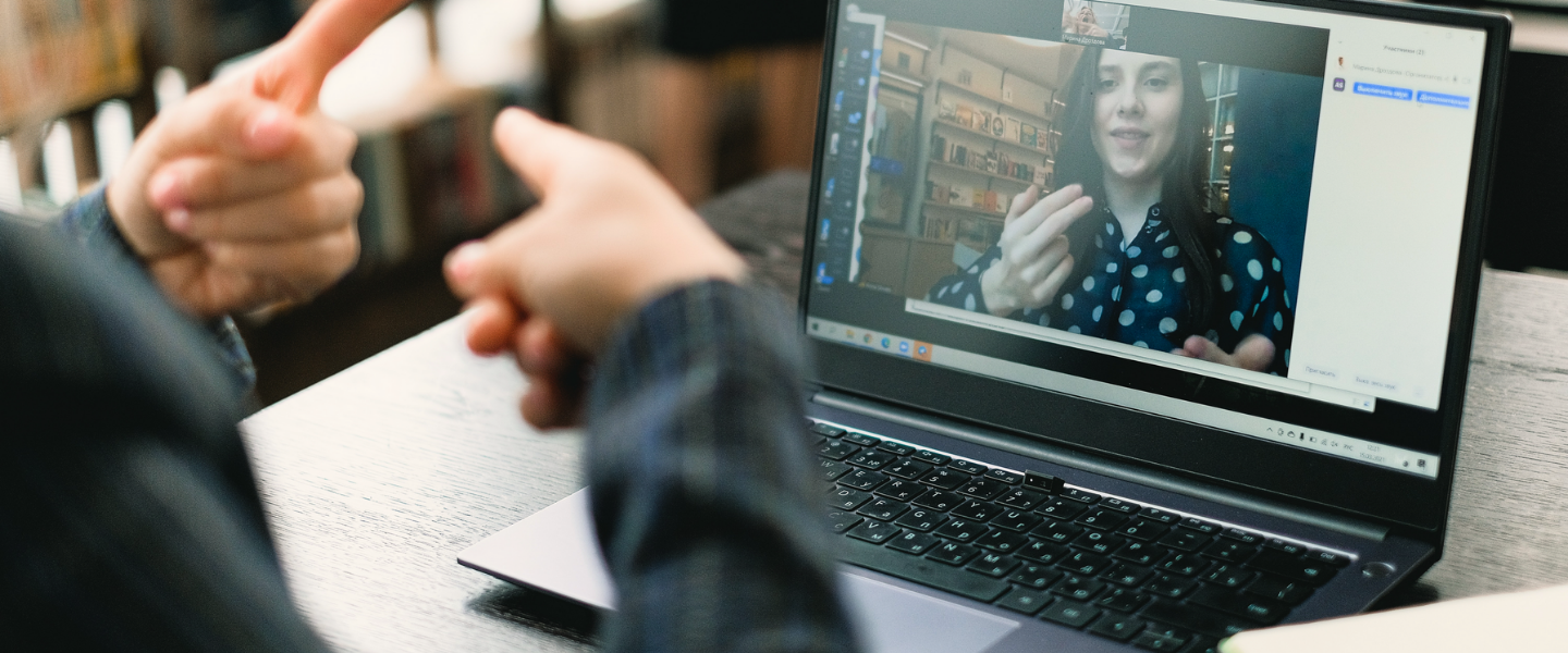 Über die Schulter einer nicht zu identifizierenden Person sieht man dessen Hände mit ausgestreckten Zeigefingern. Vor ihr steht ein Laptop. Sie unterhält sich in Gebärdensprache mit einer weiblich gelesenen Person auf dem Bildschirm des Laptops.