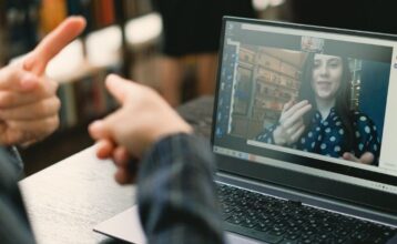 Über die Schulter einer nicht zu identifizierenden Person sieht man dessen Hände mit ausgestreckten Zeigefingern. Vor ihr steht ein Laptop. Sie unterhält sich in Gebärdensprache mit einer weiblich gelesenen Person auf dem Bildschirm des Laptops.