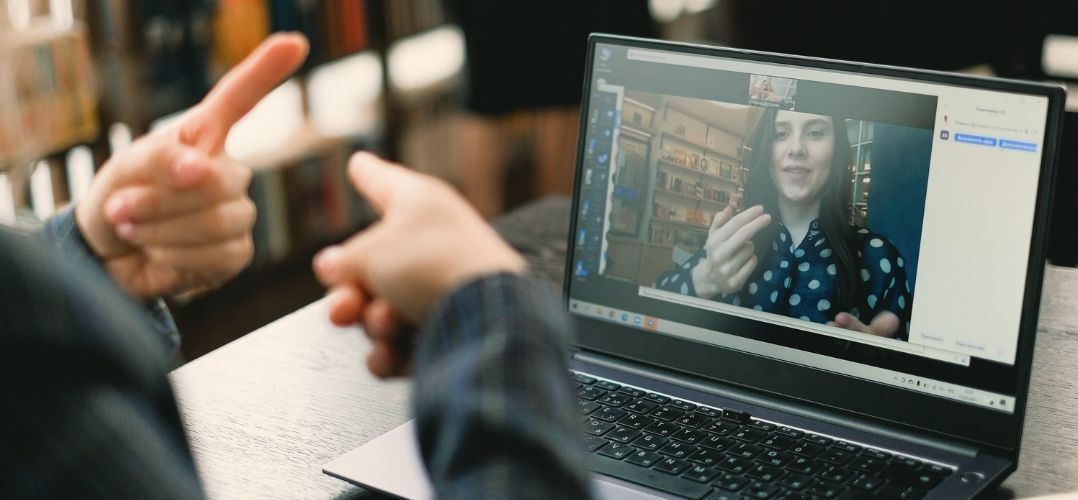 Über die Schulter einer nicht zu identifizierenden Person sieht man dessen Hände mit ausgestreckten Zeigefingern. Vor ihr steht ein Laptop. Sie unterhält sich in Gebärdensprache mit einer weiblich gelesenen Person auf dem Bildschirm des Laptops.