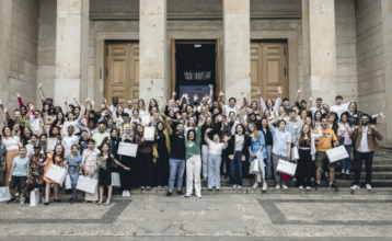 Vor einem imposanten Gebäude mit großen hellbraunen Steinsäulen und schweren Flügeltüren aus Holz stehen etwa 80 Menschen. Es ist eine diverse Gruppe mit Kindern und Erwachsenen, unterschiedlichen Hautfarben und einigen Frauen mit Hijab. Einige halten Urkunden oder Papiertüten hoch und alle winken und lachen fröhlich in die Kamera. Links und rechts stehen zwei Rollups mit der Aufschrift "Schulen gegen Diskriminierung und dem Logo der Aktion "Fair@School".