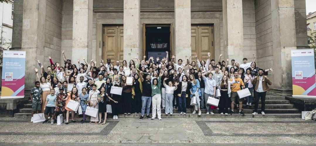 Vor einem imposanten Gebäude mit großen hellbraunen Steinsäulen und schweren Flügeltüren aus Holz stehen etwa 80 Menschen. Es ist eine diverse Gruppe mit Kindern und Erwachsenen, unterschiedlichen Hautfarben und einigen Frauen mit Hijab. Einige halten Urkunden oder Papiertüten hoch und alle winken und lachen fröhlich in die Kamera. Links und rechts stehen zwei Rollups mit der Aufschrift "Schulen gegen Diskriminierung und dem Logo der Aktion "Fair@School".