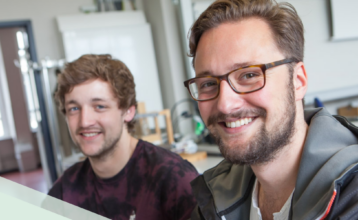 Zwei junge Männer sitzen in einem Klassenraum am Computer und lächeln in die Kamera. Der vordere ist im Fokus. Er at braune kurze Haare, einen Bart und trägt eine Brille.