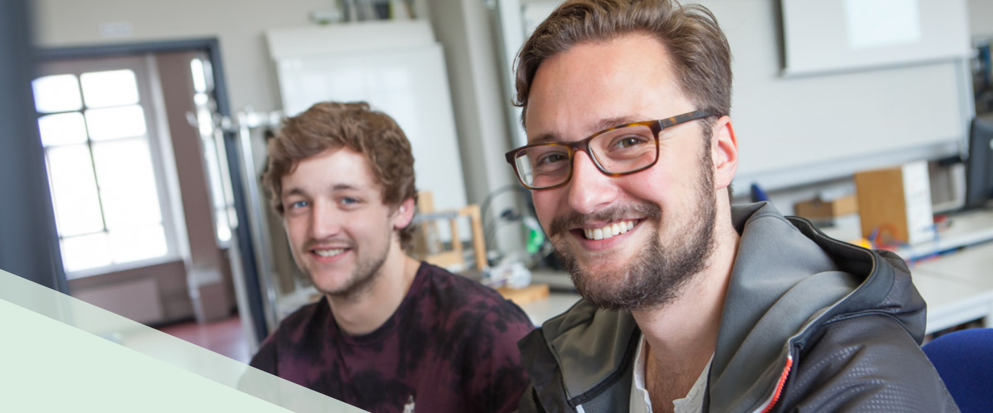 Zwei junge Männer sitzen in einem Klassenraum am Computer und lächeln in die Kamera. Der vordere ist im Fokus. Er at braune kurze Haare, einen Bart und trägt eine Brille.