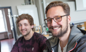 Zwei junge Männer sitzen in einem Klassenraum am Computer und lächeln in die Kamera. Der vordere ist im Fokus. Er at braune kurze Haare, einen Bart und trägt eine Brille.