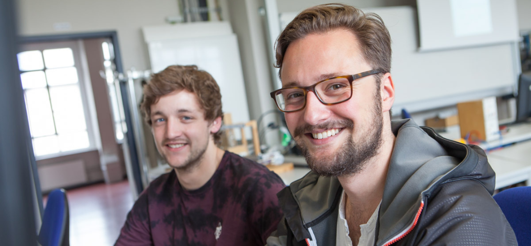 Zwei junge Männer sitzen in einem Klassenraum am Computer und lächeln in die Kamera. Der vordere ist im Fokus. Er at braune kurze Haare, einen Bart und trägt eine Brille.