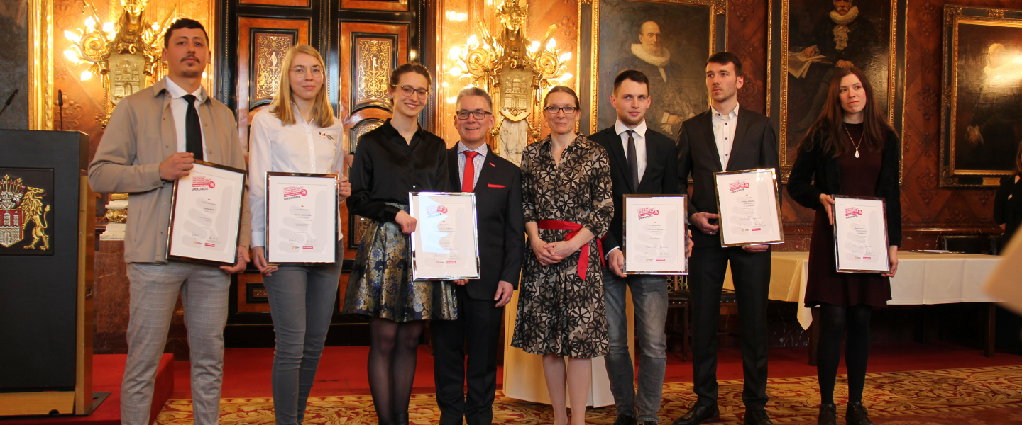 Drei junge Männer und drei junge Frauen stehen mit Urkunden in der Hand in einer Reihe. In Ihrer Mitte stehen der Präsident der Handwerkskammer, Hjalmar Stemmann und die Senatorin für Schule und Berufsbildung Ksenija Bekeris. Der Hintergrund ist der prunkvolle Bürgermeistersaal im Hamburger Rathaus.